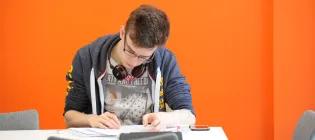 A student sits writing in front of an orange wall
